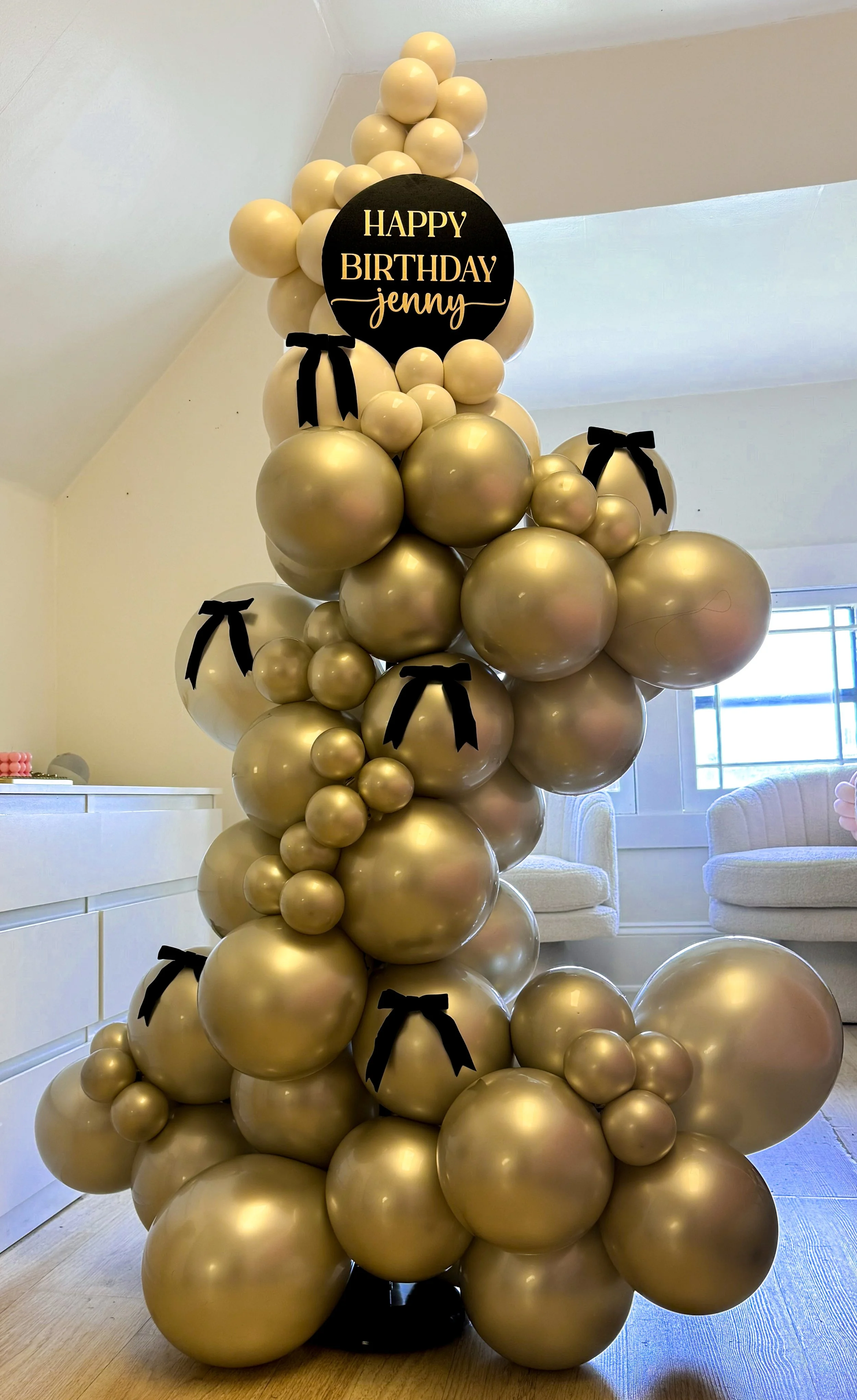 Golden balloon arrangement with a black circular sign that says 'Happy Birthday Jenny', decorated with black bows.
