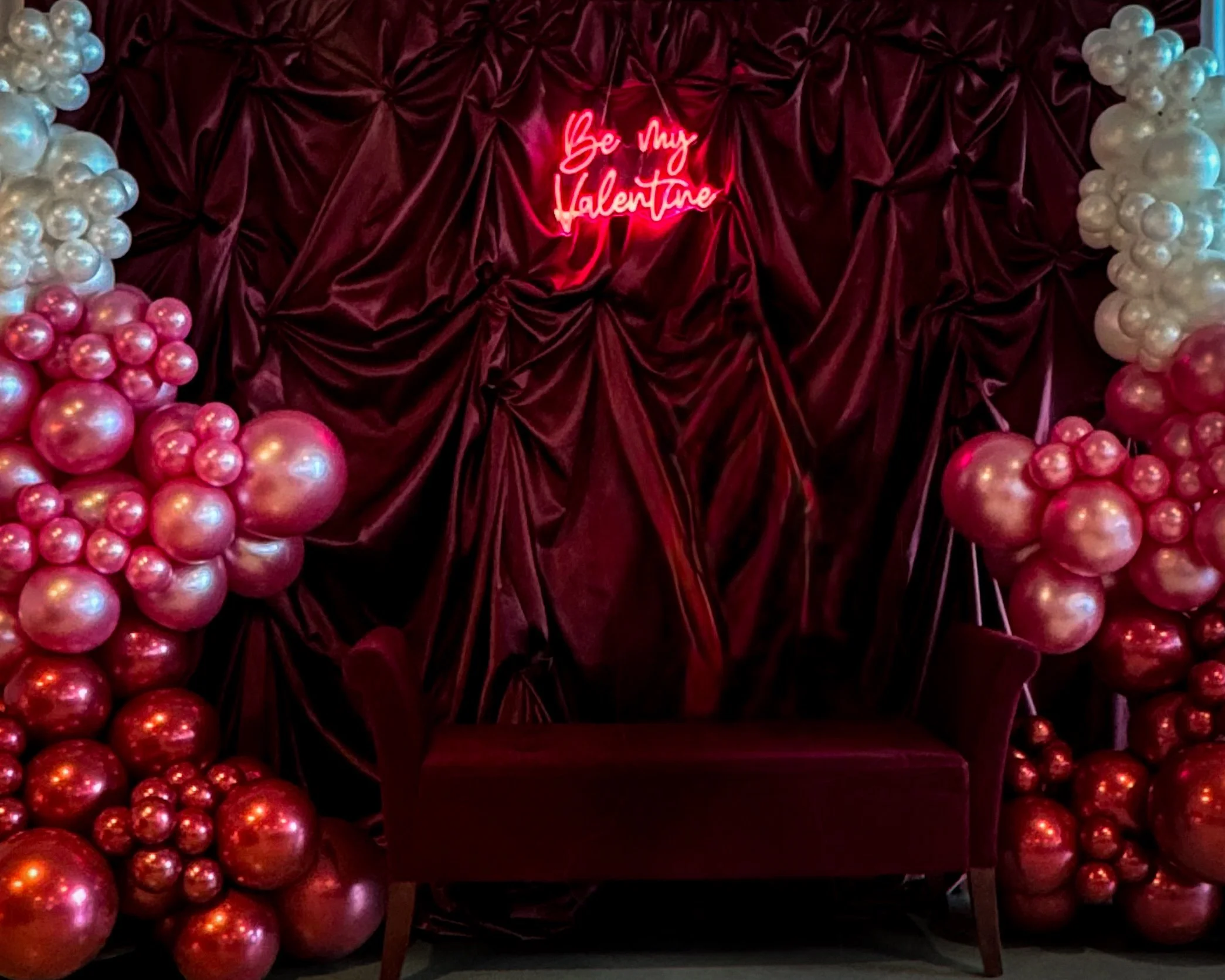 A Valentine's Day display with red velvet fabric, pink and white balloons, and a neon sign that says "Be My Valentine".