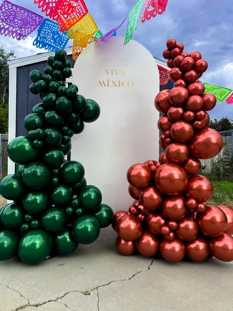 Decorative display with green and red balloons arranged in clusters, a white oval board with gold text reading 'VIVA MEXICO,' colorful papel picado banners, and a light purple unicorn horn attached to the top of the board, celebrating Mexican culture.