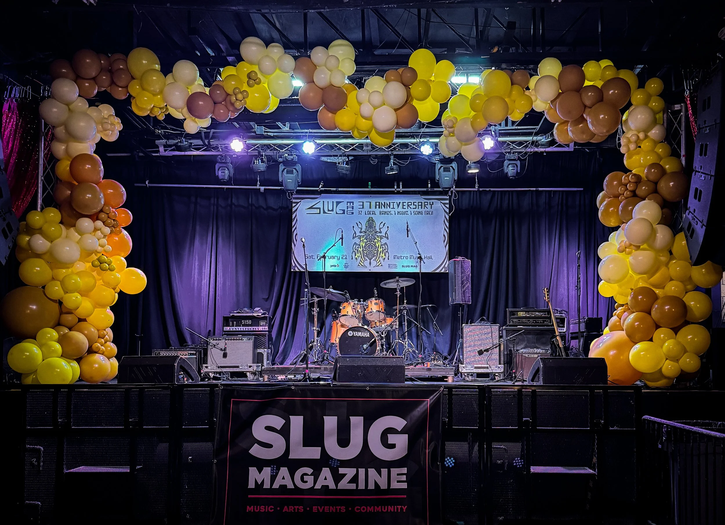 Stage decorated with yellow, cream, orange, and brown balloons arching across the top and sides. The stage features musical instruments like drums and amplifiers, with a backdrop displaying a banner celebrating the 37th anniversary of SLUG Magazine. The stage setup suggests a music or community event.