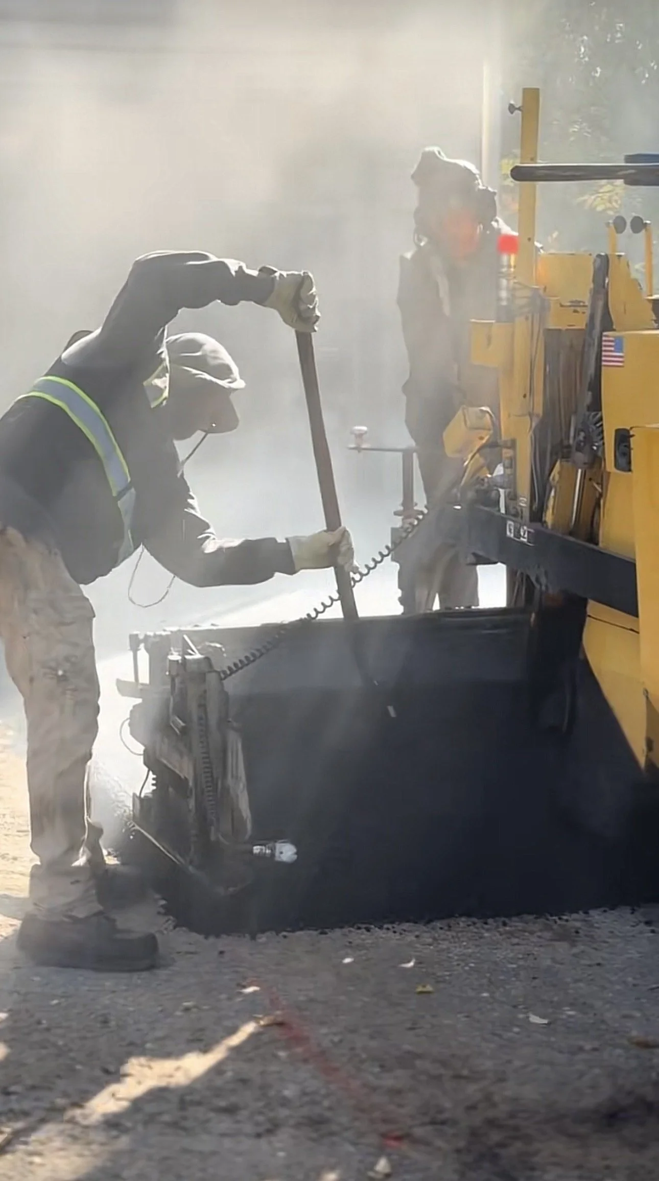 Two construction workers operating asphalt paving equipment, wearing safety gear, with dust and sunlight in the background.