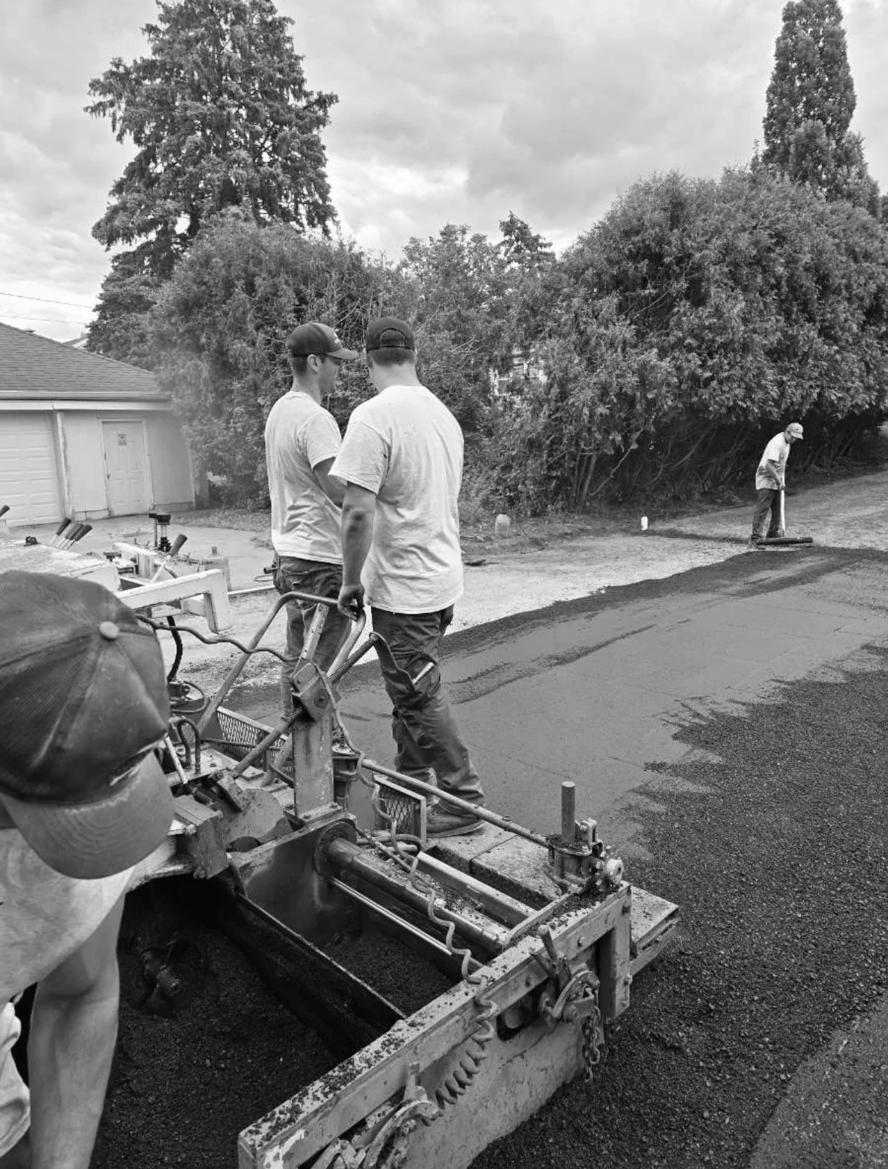 Two men stand and converse near fresh asphalt being laid on a driveway while another worker smooths the surface in the background. Trees and a garage are visible in the background.