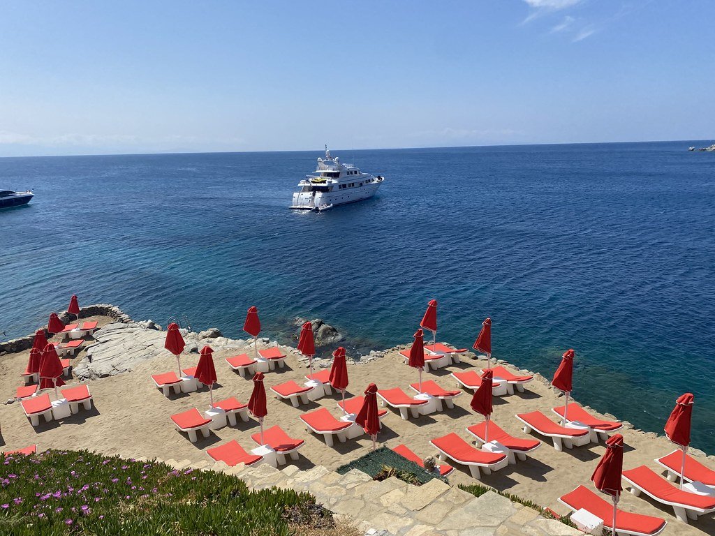 Beach with red lounge chairs and umbrellas on sand, overlooking the ocean with a large yacht and other boats.
