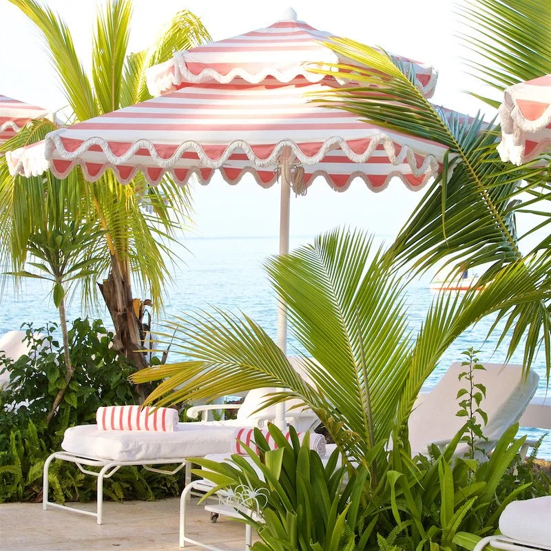 Beachside outdoor lounge area with a pink and white striped umbrella, lounge chair with matching pillows, surrounded by tropical green plants and palm trees, overlooking the ocean.