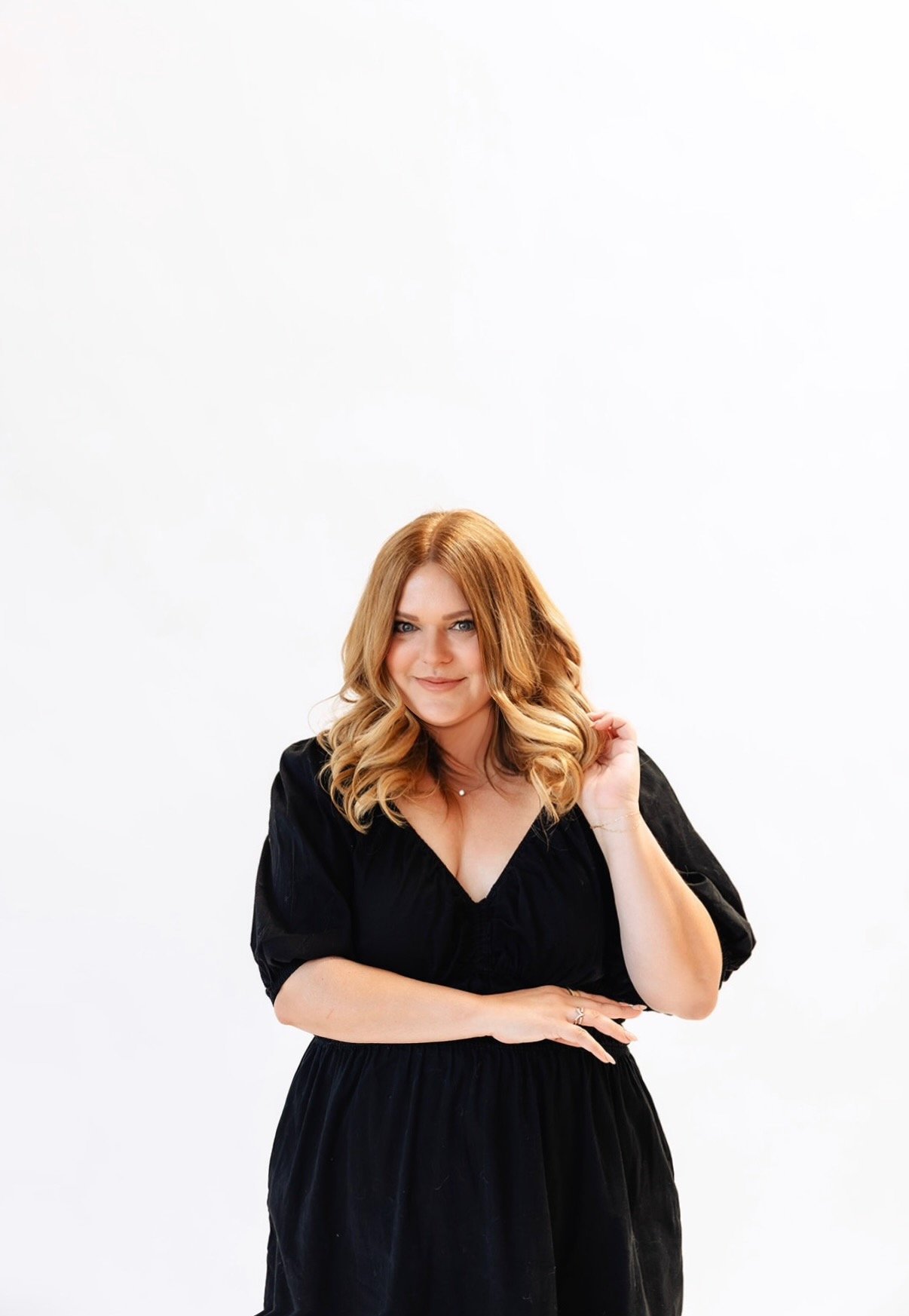 A woman with shoulder-length blonde hair, wearing a black dress, smiling and posing with one hand near her hair against a plain white background. Kelli from Let Your mind Unwind.