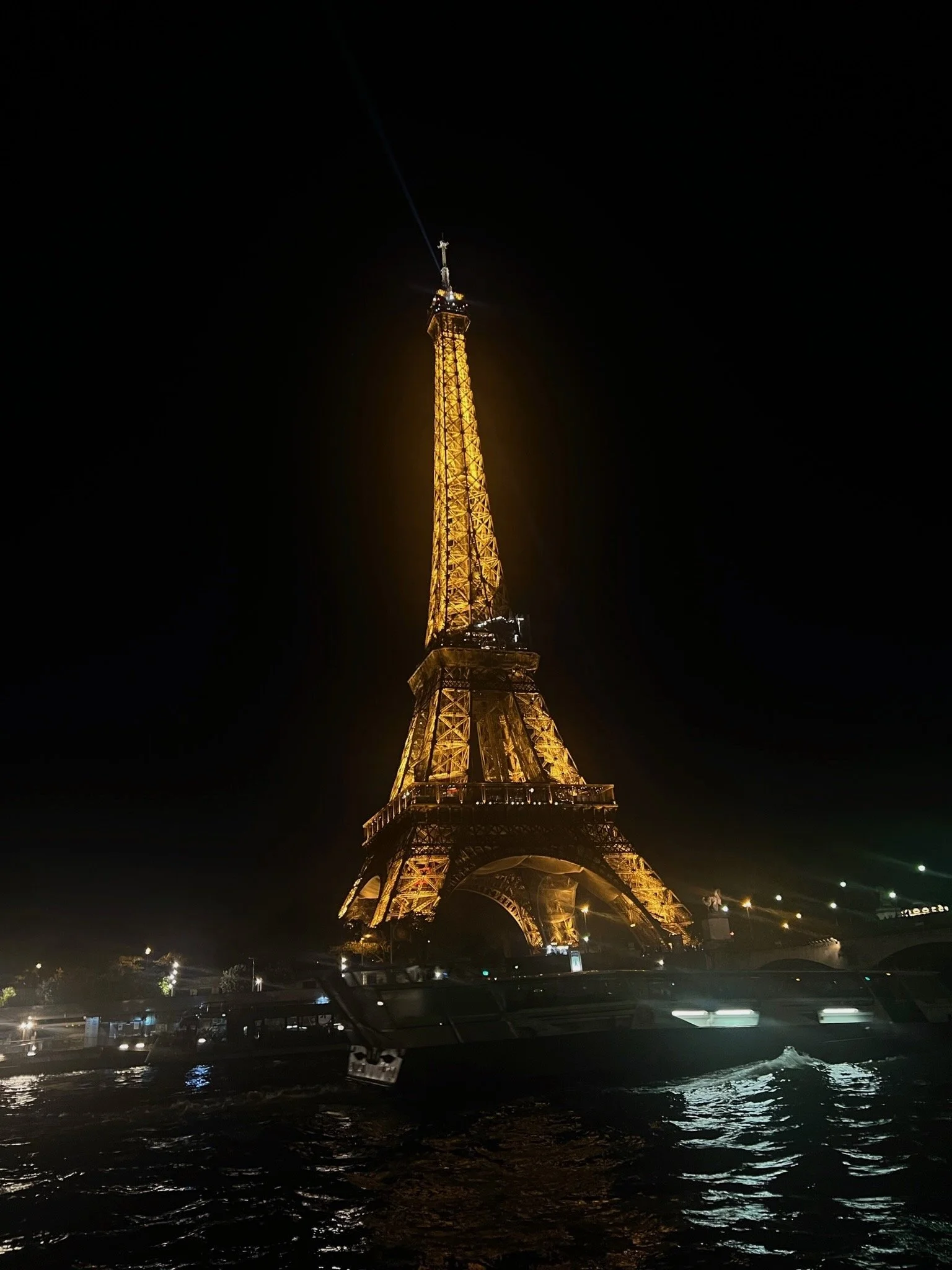 The Eiffel Tower illuminated at night, seen from the water with boats and reflections on the surface.