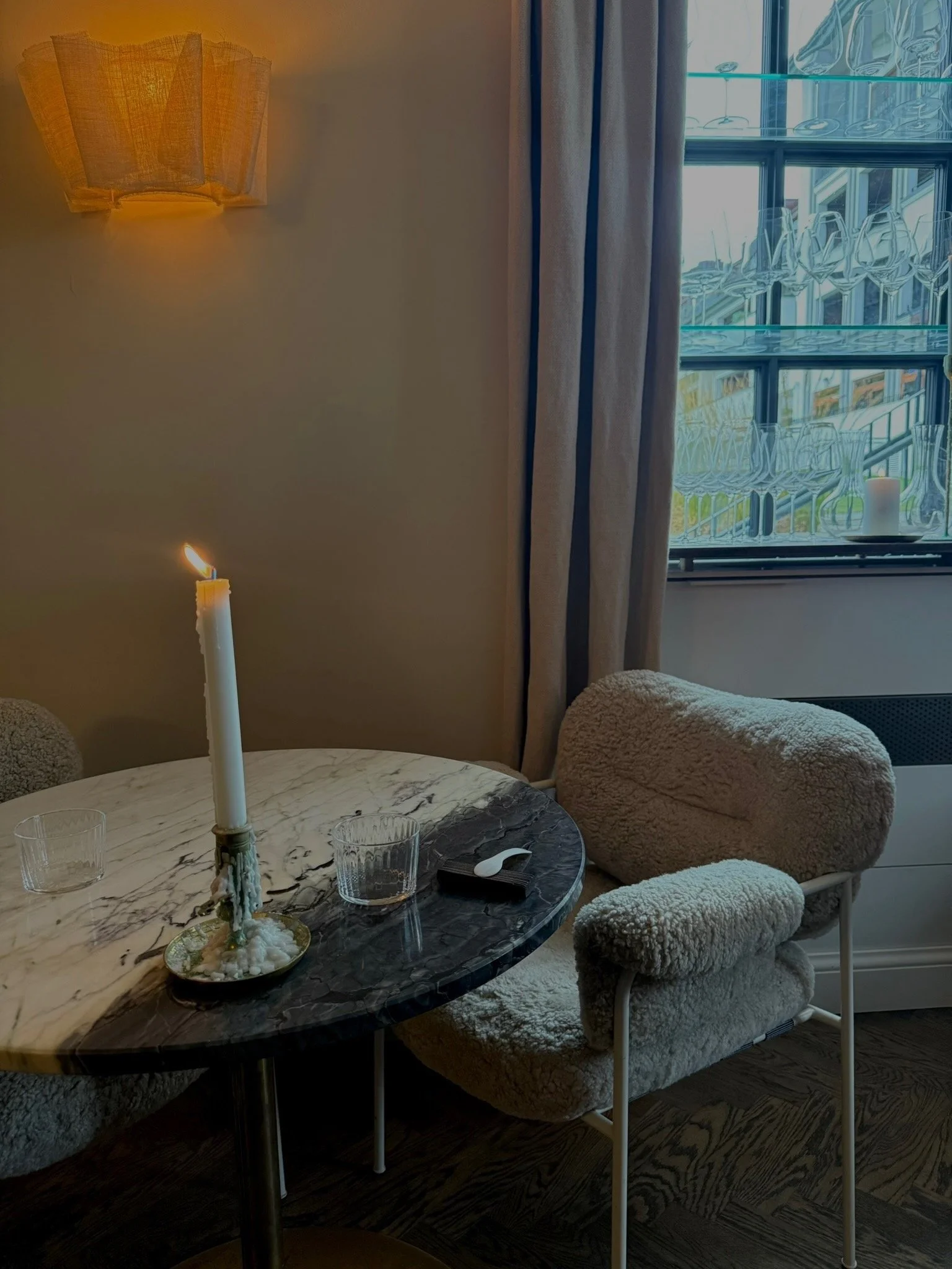 A cozy living room corner with a marble table, a lit candle on a decorative holder, two empty glasses, and plush beige chairs. A window with patterned glass and curtains is in the background.