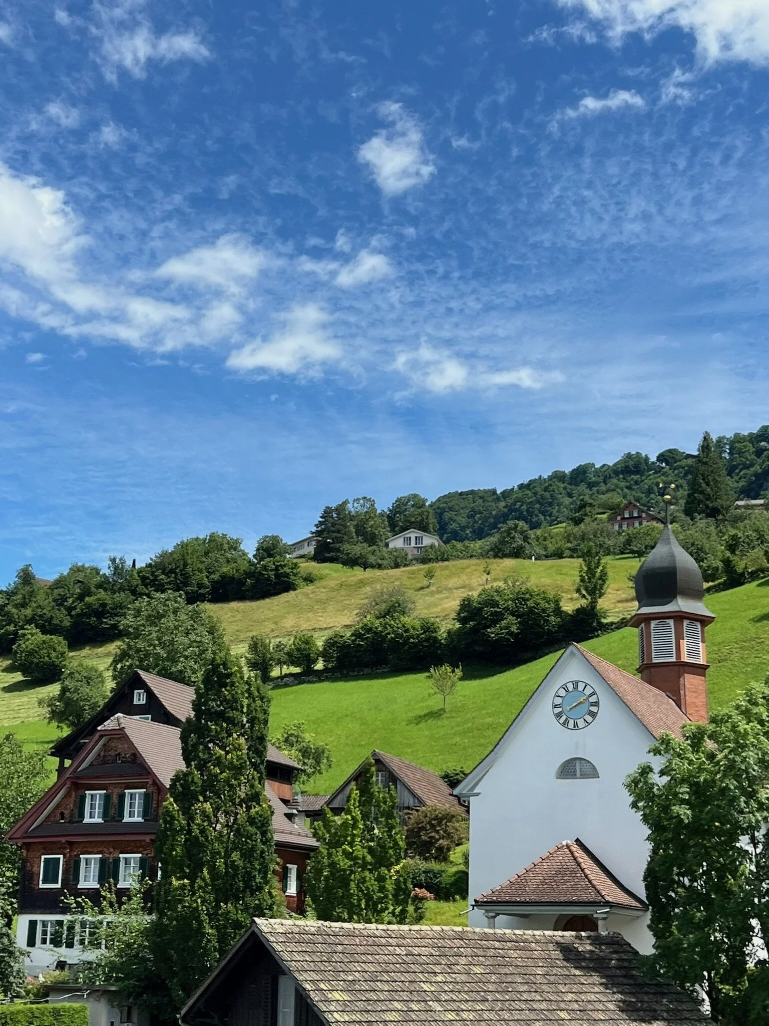 A scenic view of a small village in a hilly area with several houses and a church with a clock, surrounded by lush green trees and grass, under a blue sky with scattered clouds.