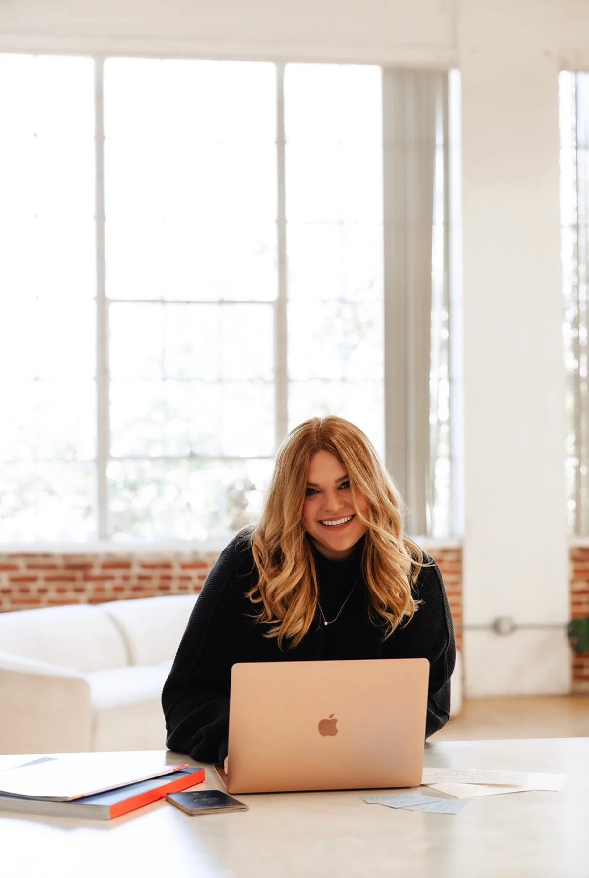 A smiling woman with long wavy hair sitting at a desk with a silver MacBook laptop, in a bright room with large windows, exposed brick wall, and white furniture. She's a travel agent booking someone's dream vacation.