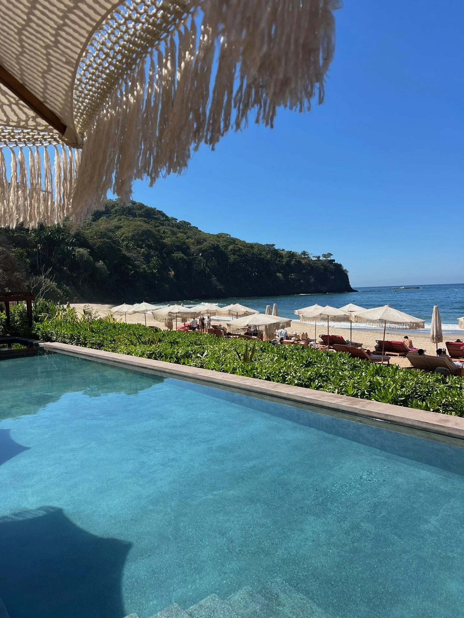 View of a beach with sun umbrellas and loungers, a cliff with trees in the background, and a clear blue sky, seen from a swimming poolside with a straw beach umbrella partially visible.