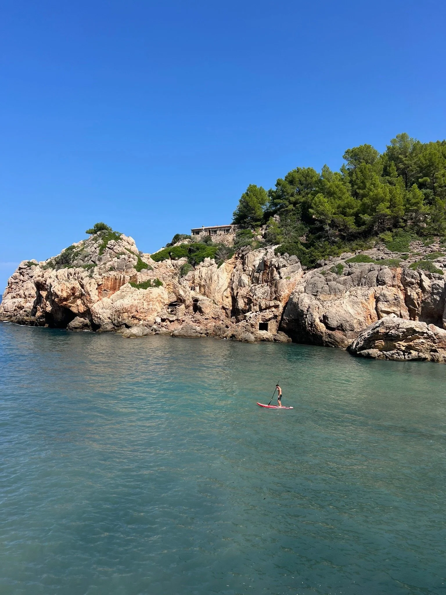 A person stand-up paddleboarding in a body of water near rocky cliffs and a hillside covered with green trees under a clear blue sky.