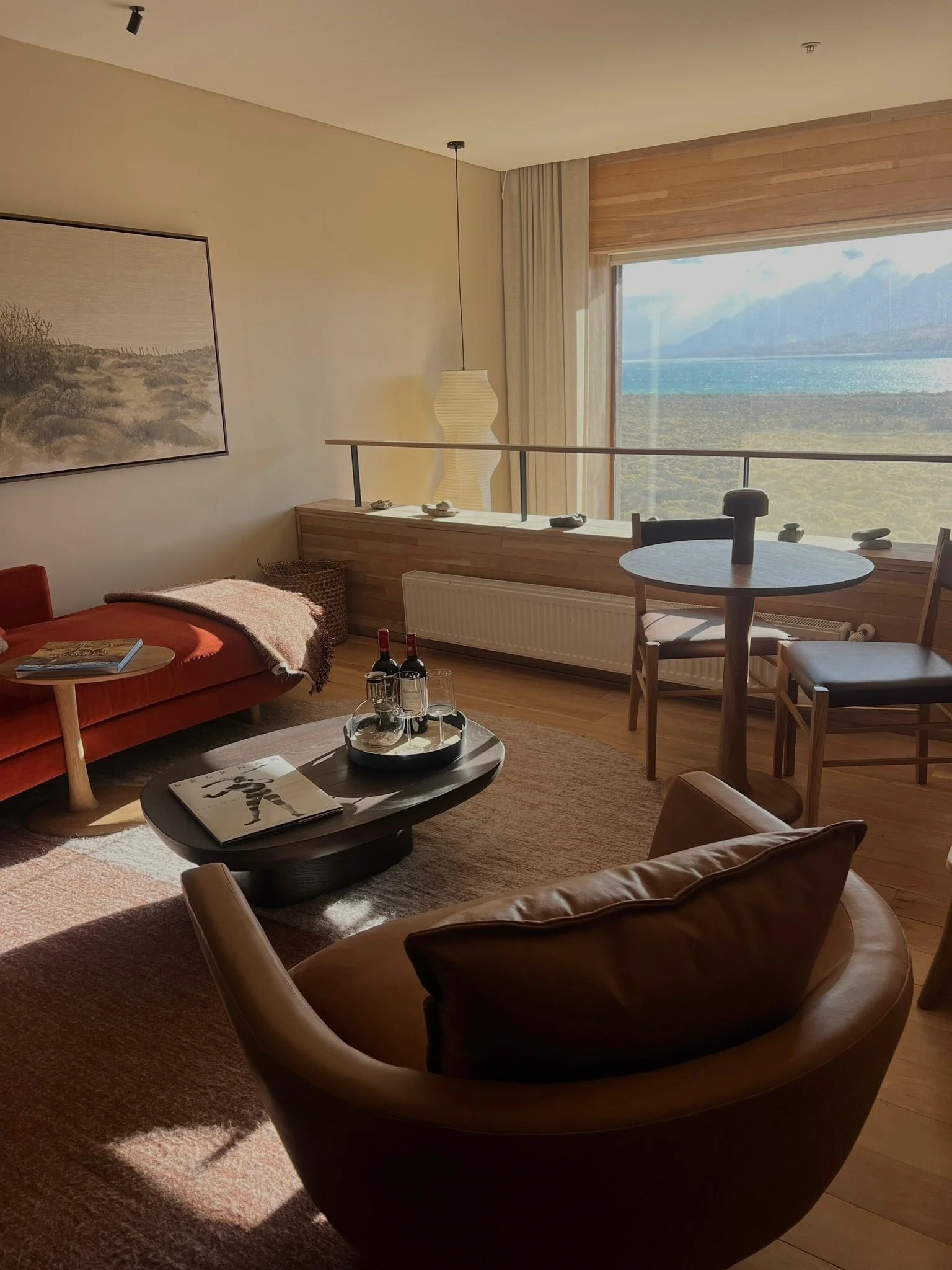 Living room with a brown leather sofa, a round coffee table with magazines and bottles, a dining table with chairs, a large window with mountains view, a framed landscape artwork, and a tall white floor lamp.