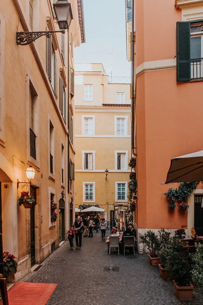 A narrow European street with colorful buildings, outdoor seating, and pedestrians.