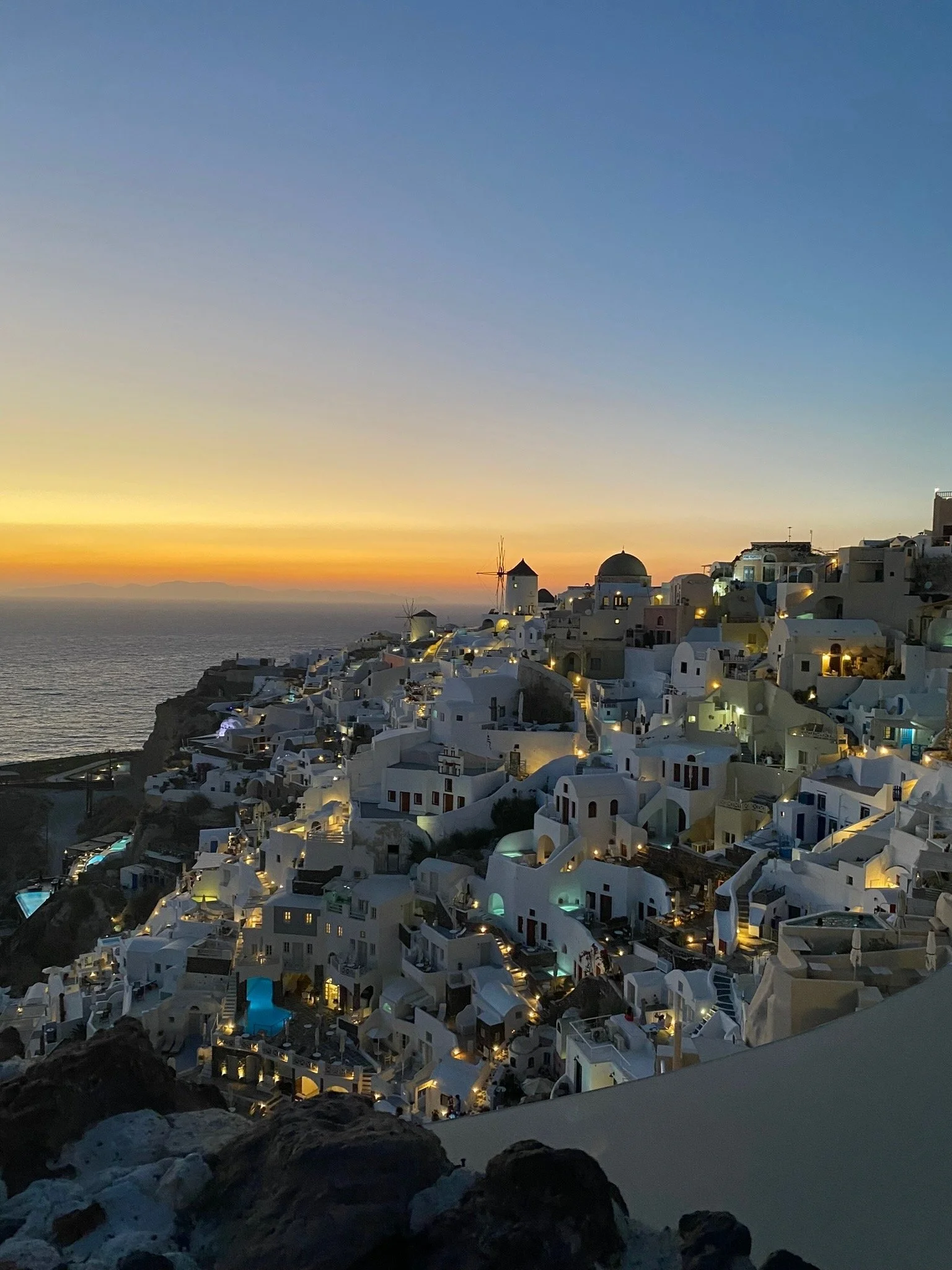 scenic view of white buildings on a cliffside at sunset, with a windmill, a domed church, and the ocean in the background, typical of Santorini, Greece