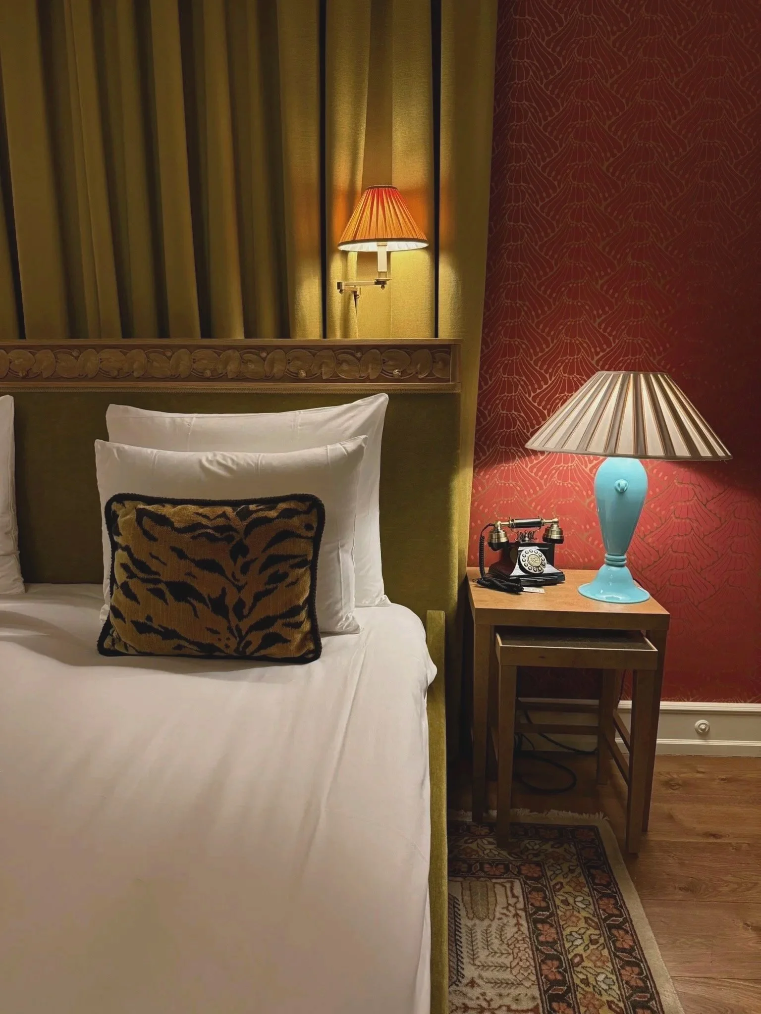 A cozy hotel room corner with a bed, decorative pillow, two table lamps with shades, a vintage rotary phone, and a side table. The room has contrasting wallpapered walls, one with yellow curtains and the other with reddish patterned wallpaper. A patt