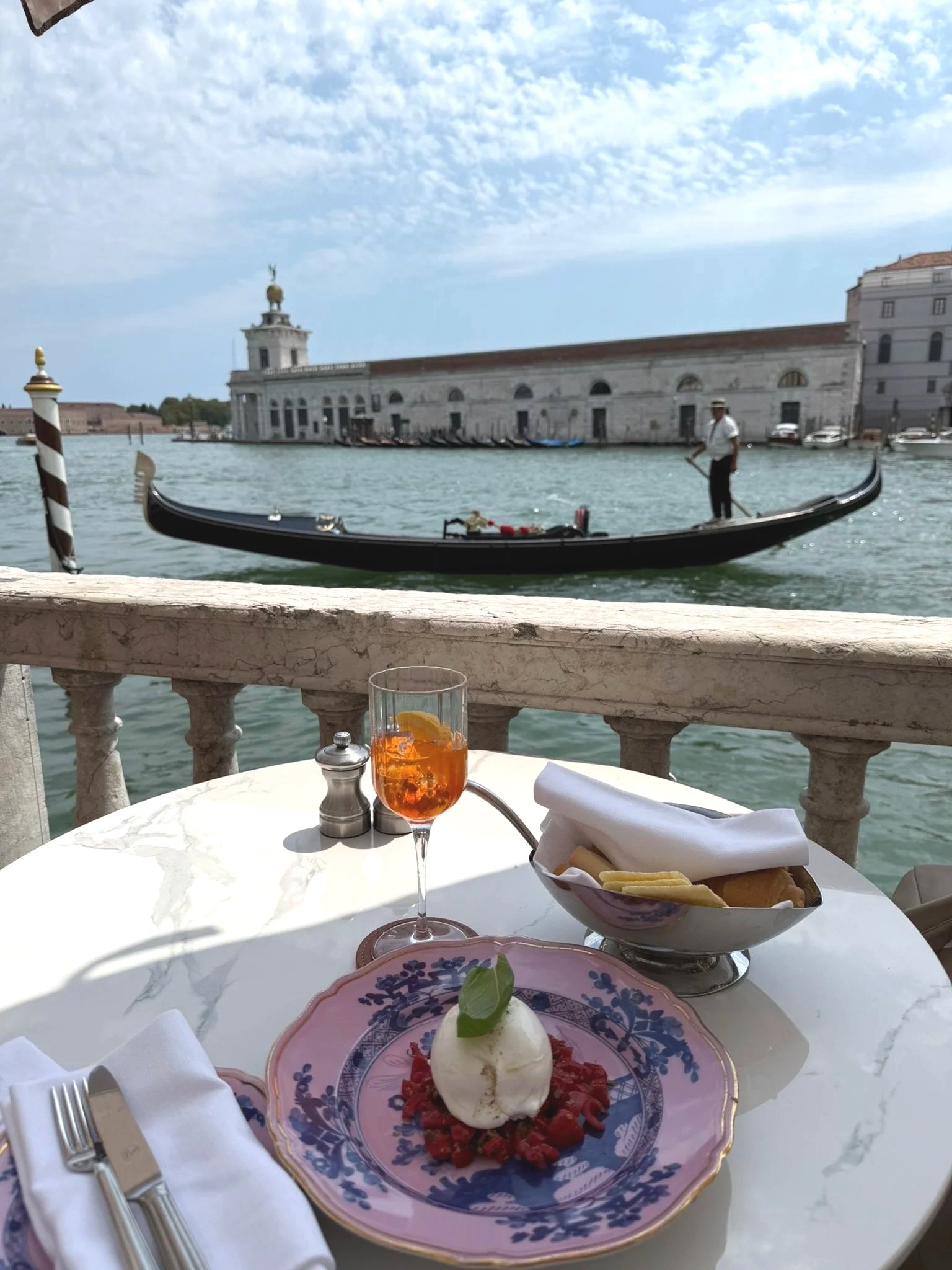 An outdoor dining table with a dessert of strawberries, whipped cream, and a mint leaf, along with a drink and salad, overlooking a canal with gondolas and a historic building, in Venice, Italy.