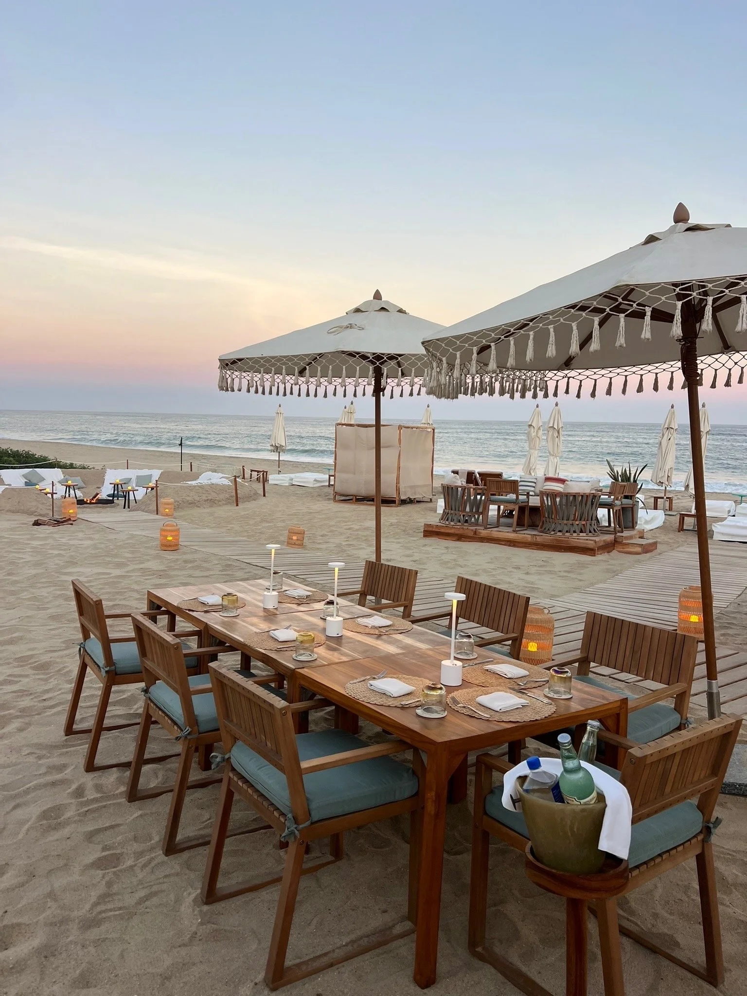 Beautyful beach restaurant setup with wooden tables, chairs, large white umbrellas with tassels, and the ocean in the background during sunset.