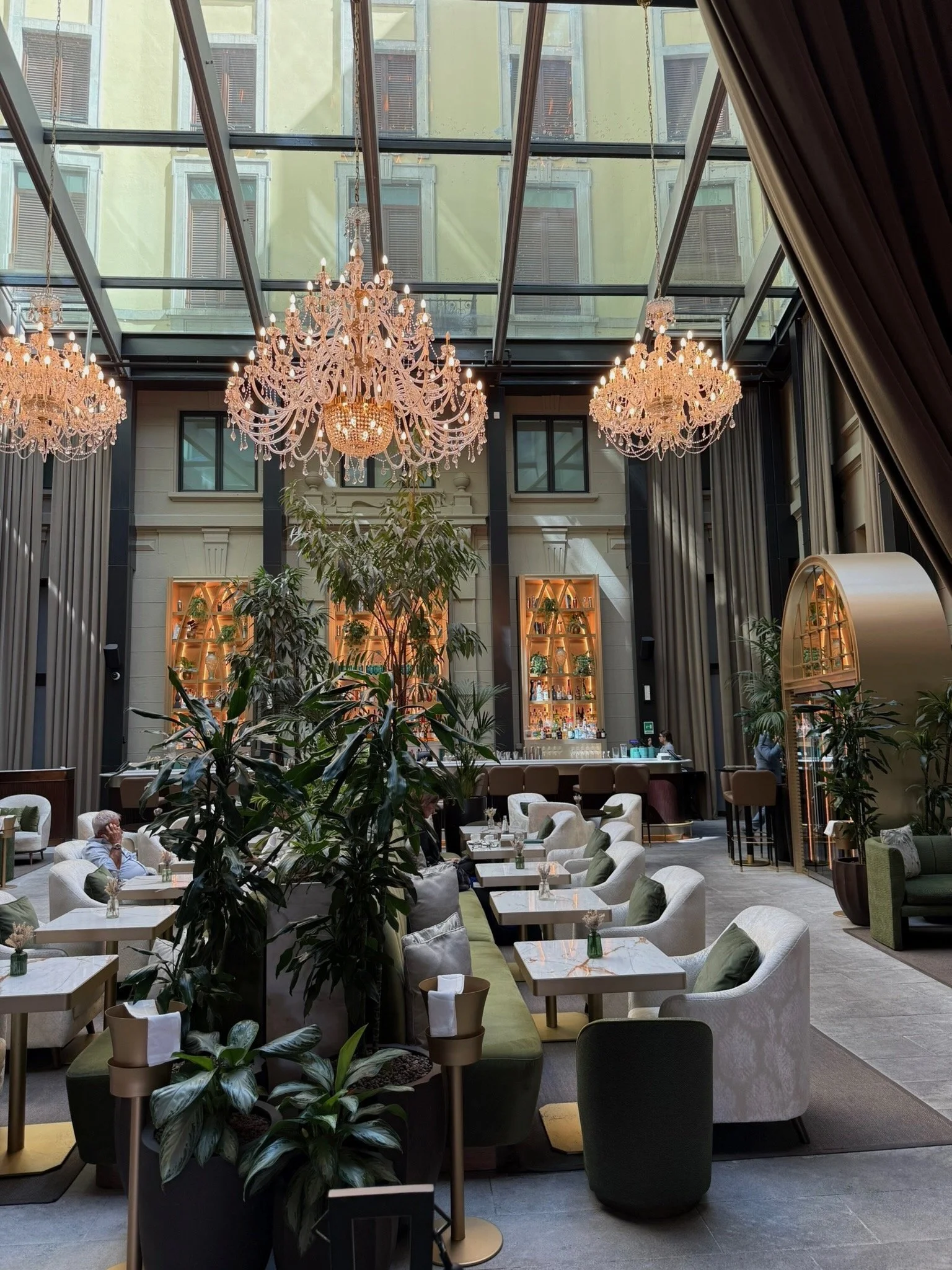 Interior of a modern restaurant or hotel lounge with large chandeliers hanging from a glass ceiling, greenery, and a bar area in the background. Seating includes white and green upholstered chairs and tables.