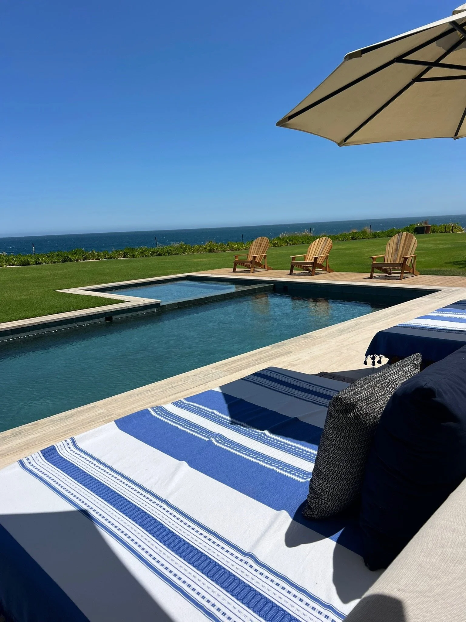 View of a poolside area with striped rug, pillows, and outdoor seating, overlooking a grassy lawn and ocean under a clear blue sky with a large umbrella in the corner.