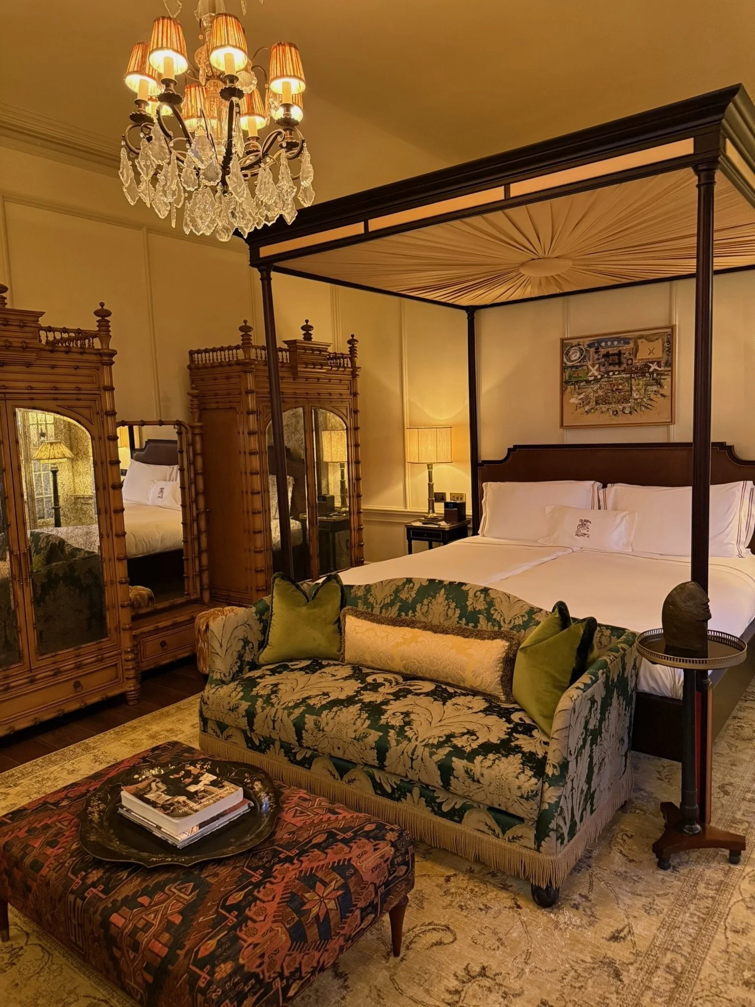 A luxurious hotel room featuring a four-poster bed with white bedding, a vintage wooden wardrobe, a plush floral-patterned sofa with green and gold pillows, a chandelier hanging from the ceiling, and a richly patterned ottoman with books on it. The room is warmly lit with table lamps and has a decorative painting on the wall.