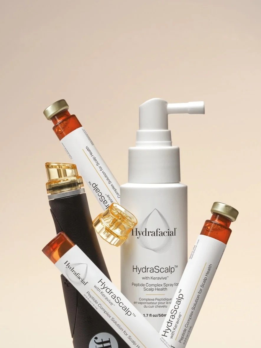 Varios frascos y un spray de un producto para la salud del cuero cabelludo, llamado HydraScalp, en diferentes colores y tamaños, sobre fondo neutro.