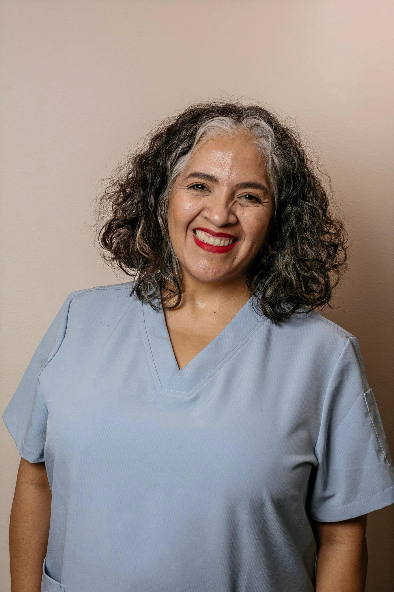 Mujer sonriendo con pelo rizado y canoso, vestida con uniforme de color azul claro contra una pared beige.