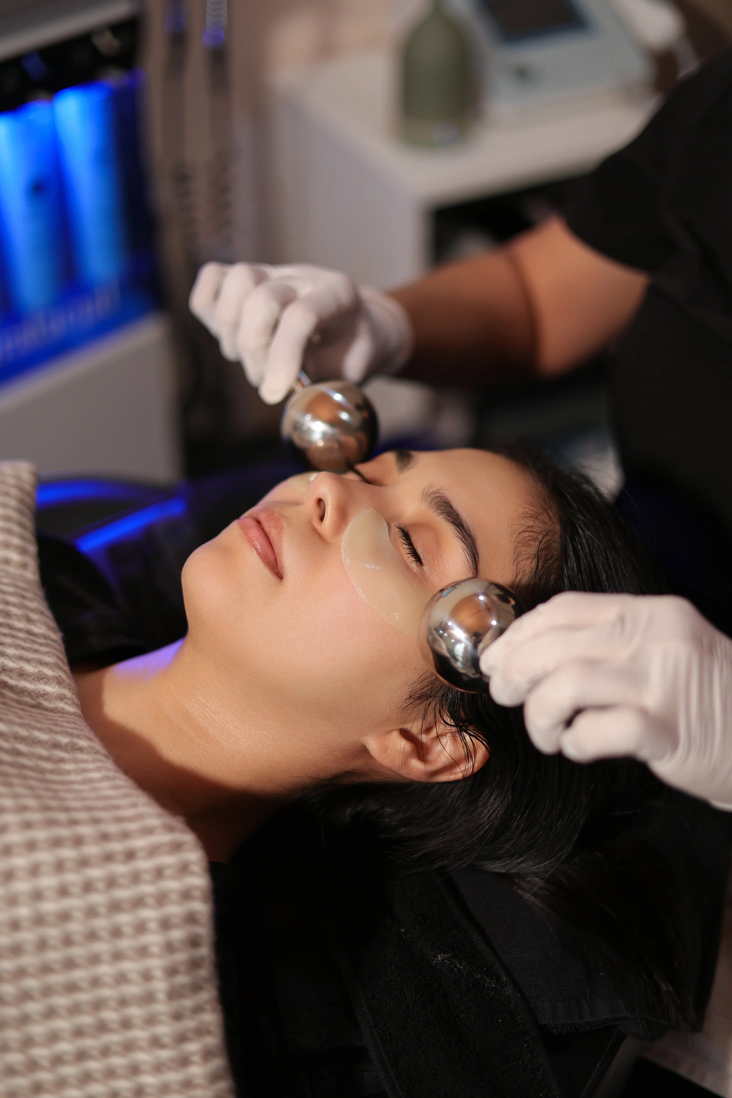 Una mujer recibiendo un tratamiento facial en un spa o clínica estética, con terapeutas usando bolas metálicas en su rostro.