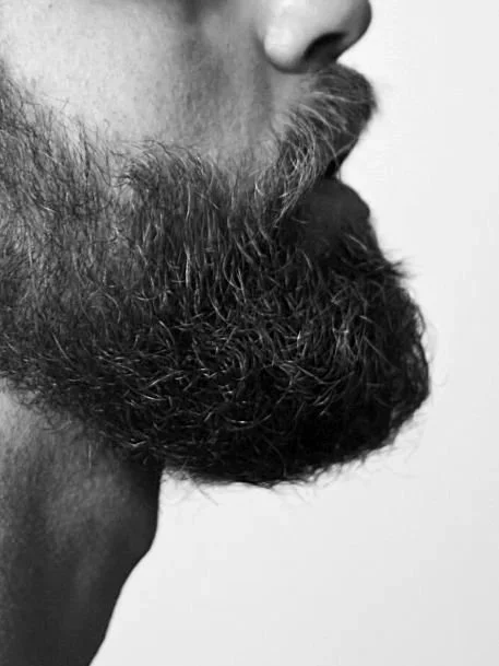 Primer plano de una barba negra y rizada, con un poco de bigote, en un fondo gris.