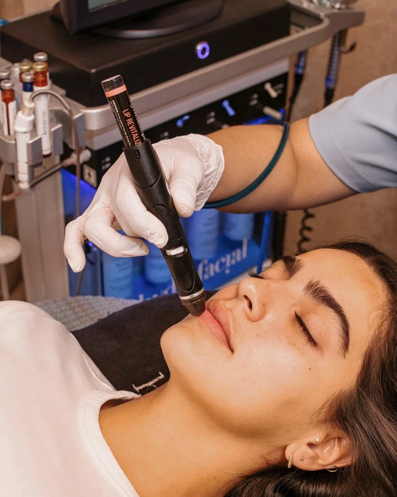 Una mujer recostada con ojos cerrados recibe un tratamiento facial con un dispositivo eléctrico en el rostro, en un ambiente de clínica estética.