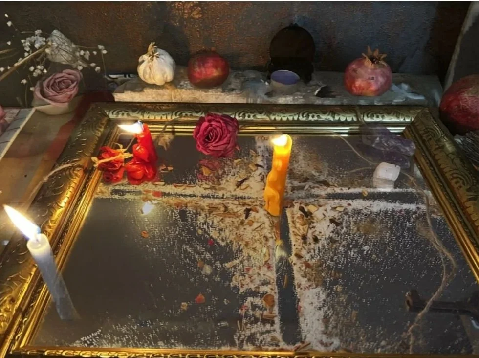 A mirror on a table surrounded by candles, roses, pomegranates, and other objects, some of which are burnt or charred.