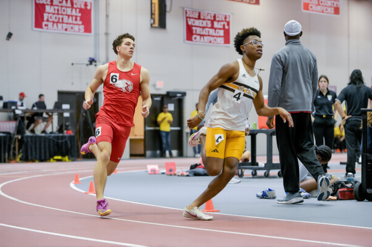 Two male athletes running on an indoor track during a race, with officials and spectators in the background, and banners hanging on the wall.