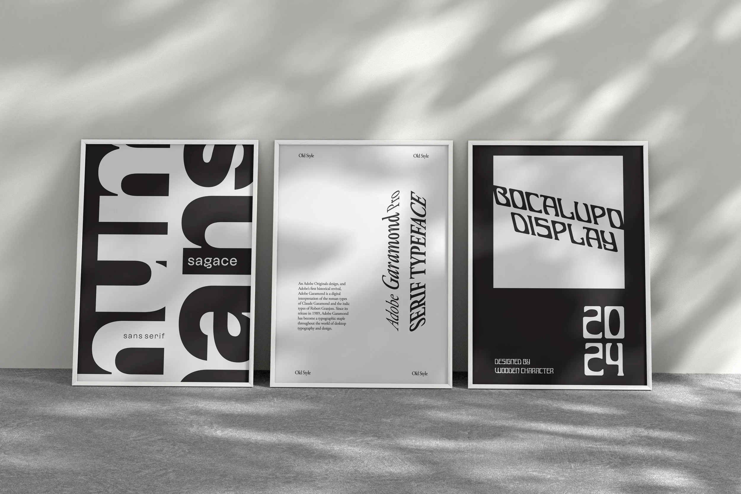 Three framed posters featuring bold, black and white text and graphics, standing on a concrete floor against a light-colored wall with shadows.