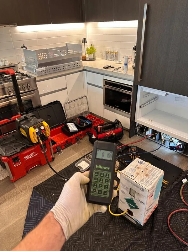 Person wearing a glove holding a Megger insulation resistance tester in a kitchen under repair, with tools and equipment on a workbench, open cabinet, and kitchen appliances visible.