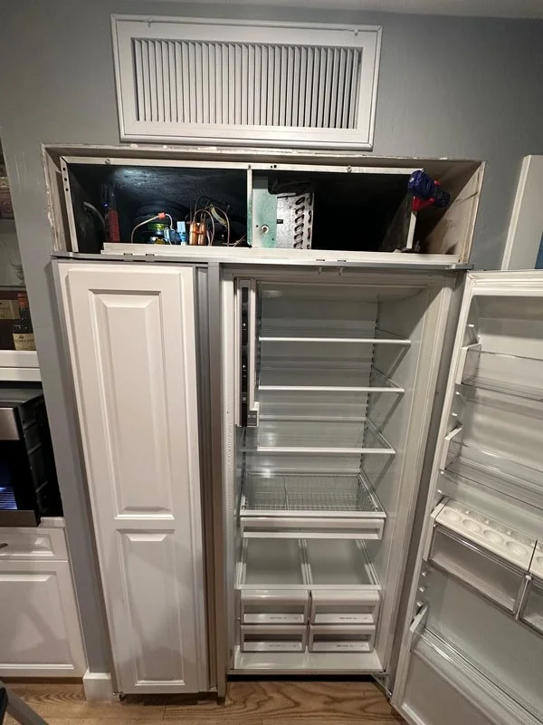 Open refrigerator with empty shelves and drawers, adjacent to a built-in cabinet, with an accessible top compartment containing tools and electronic components.