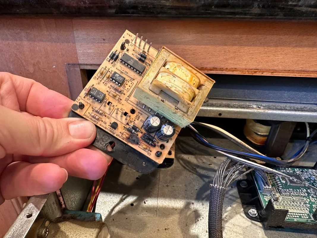 Person holding a circuit board with a transformer, capacitors, and other electronic components inside a machine or device.