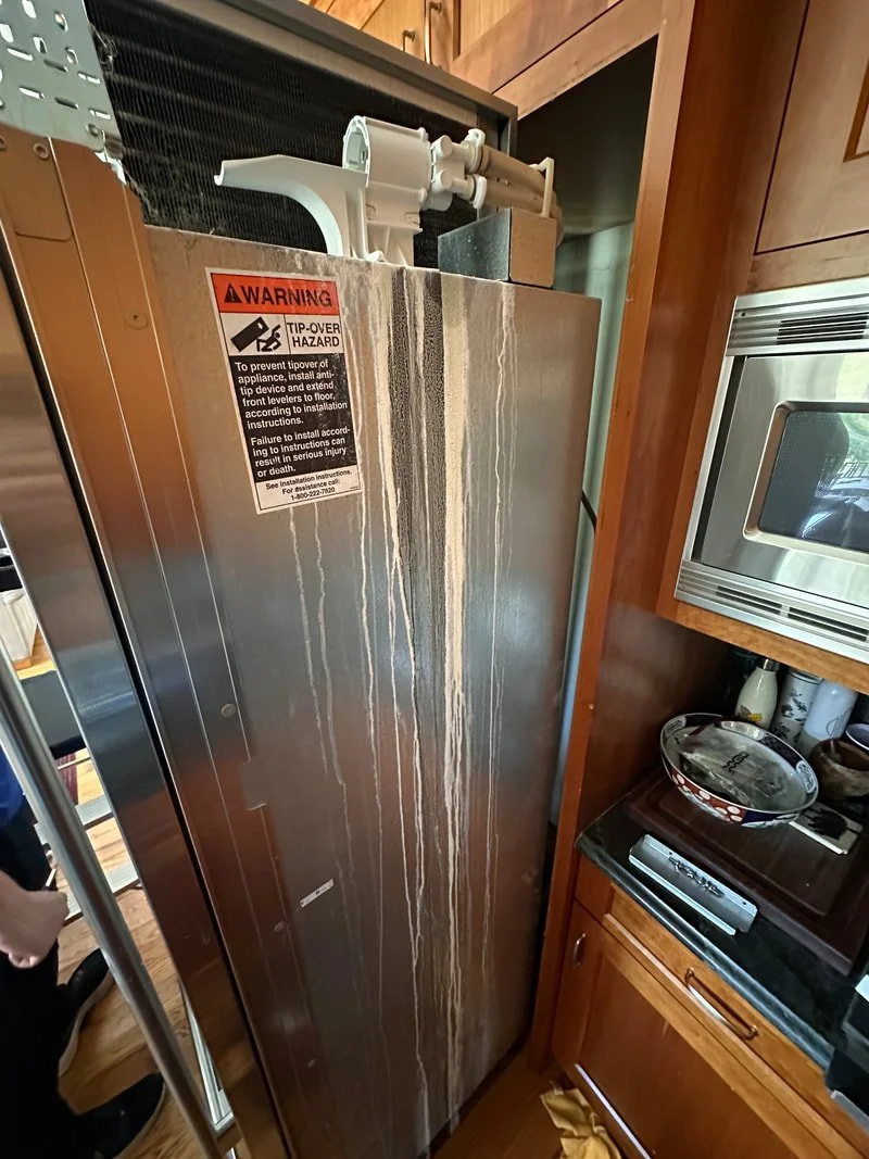 A stainless steel refrigerator with white streaks and an orange warning sticker on the front, situated in a wooden kitchen, next to a microwave and countertop.