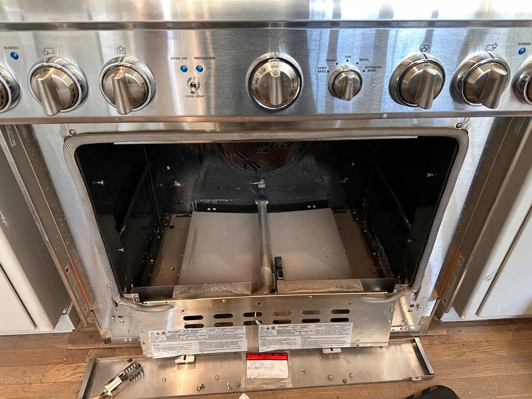 Empty stainless steel oven with its door removed, exposing the interior, placed on a wooden floor.