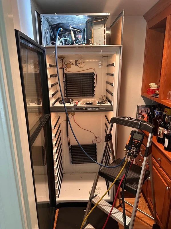 Open freezer with electrical components and tools inside, connected to testing equipment on a chair in a kitchen.