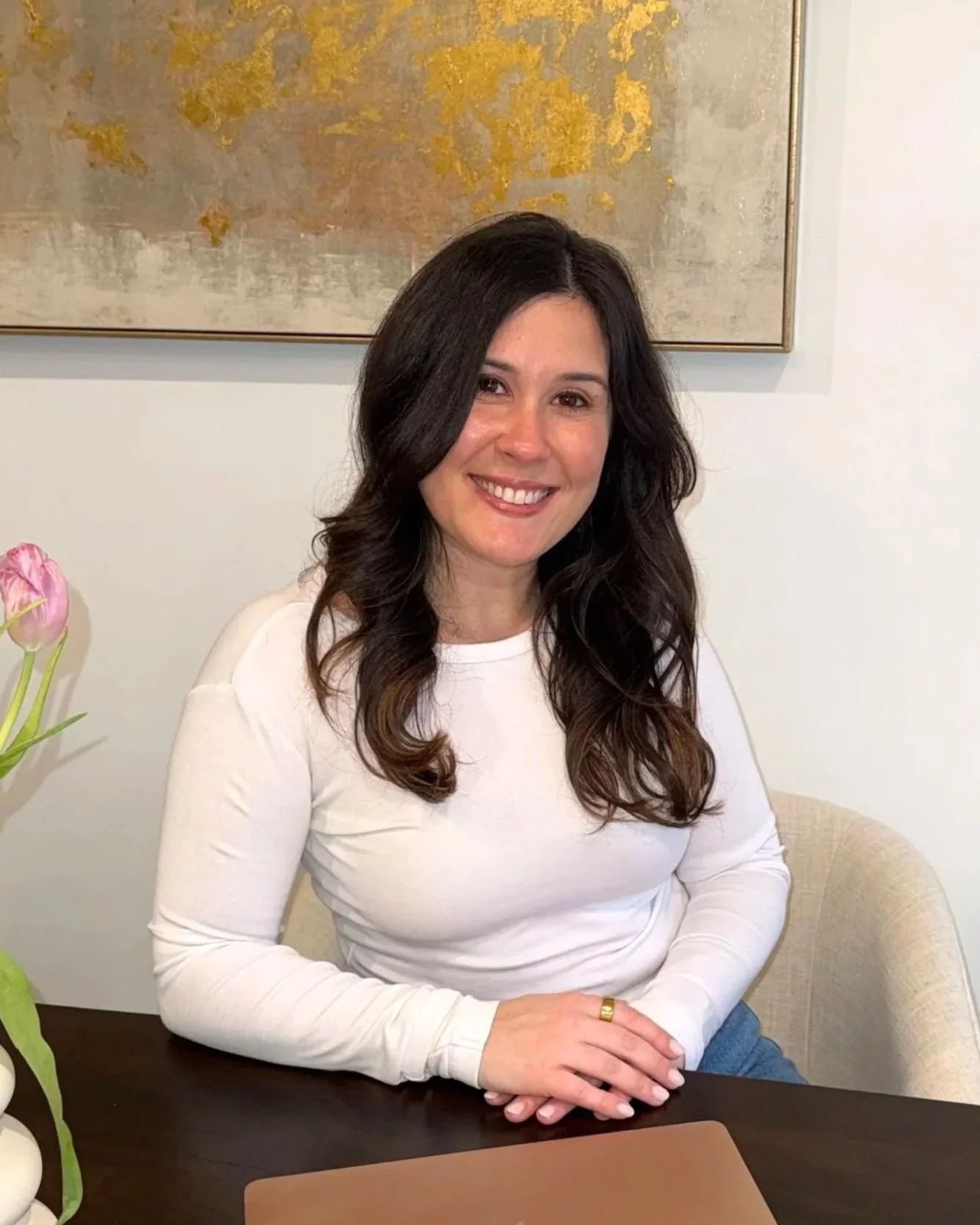 A woman with long dark wavy hair, wearing a white long-sleeve shirt, sits at a dark wooden table, smiling at the camera. There is a beige chair, a pink flower on the table, a framed abstract painting with gold accents on the wall behind her.