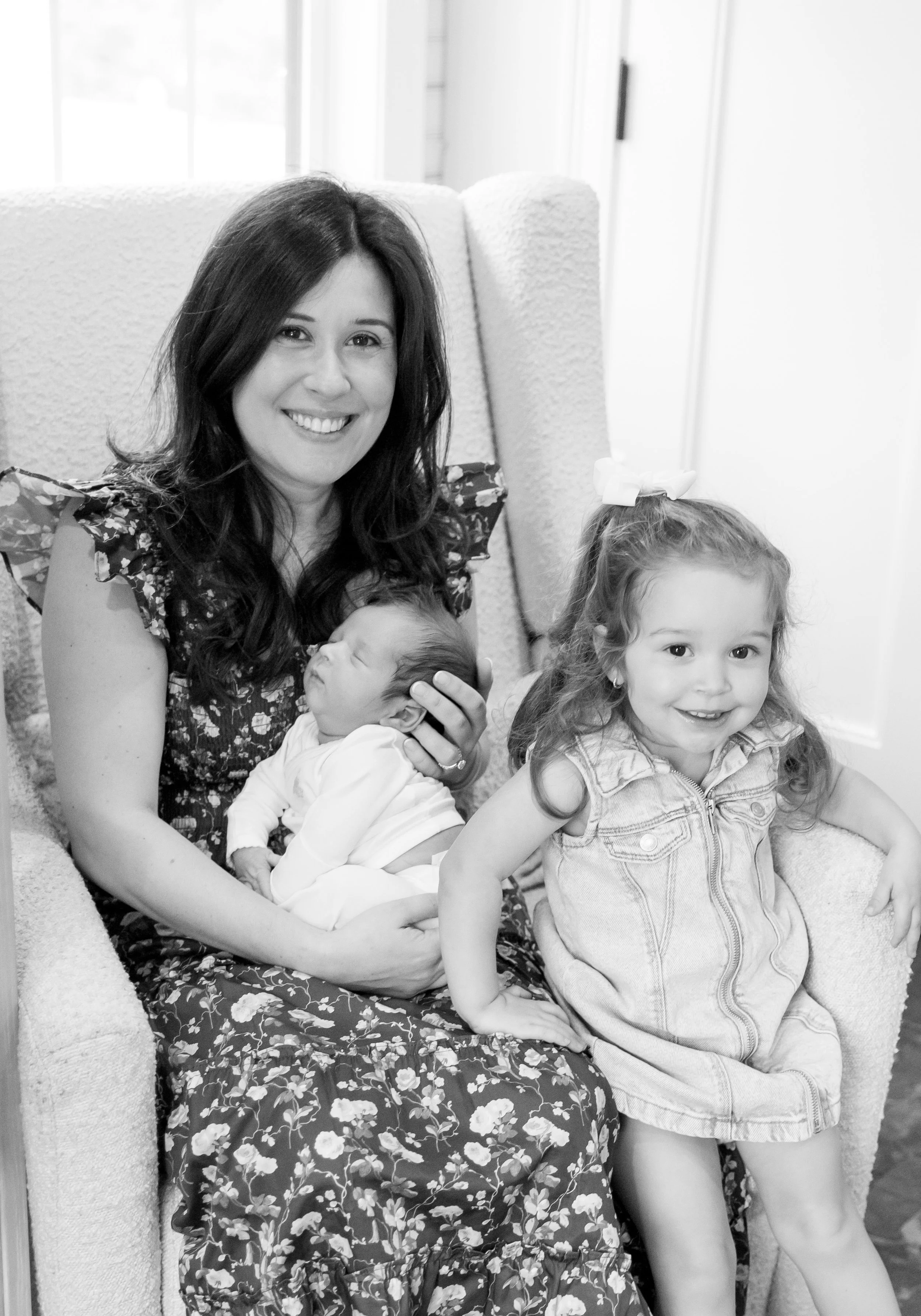 A woman sitting in a chair holding a newborn baby, with a young girl sitting next to her, all smiling.