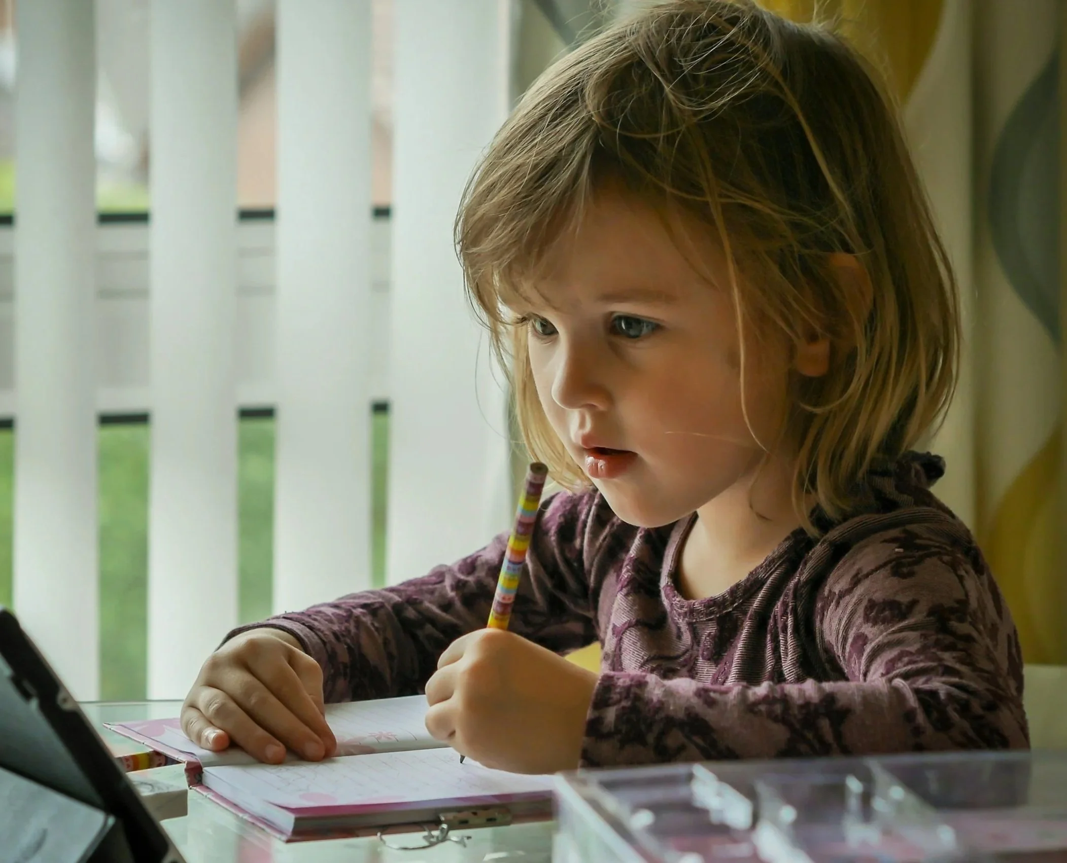 A young girl with shoulder-length brown hair and blue eyes is sitting at a table, looking intently at a tablet on her left while holding a colorful striped pencil in her right hand. She has an open notebook in front of her and appears to be engaged in her work near a window with vertical blinds.