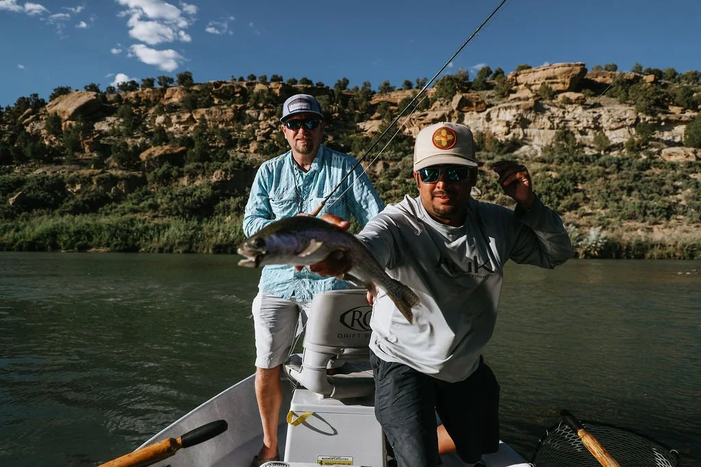 Two men on a boat in a river with rocky hills in the background; one man in the foreground showing off a fish he caught, holding it with one hand while wearing sunglasses and a cap, the other man in the background holding a fishing rod.