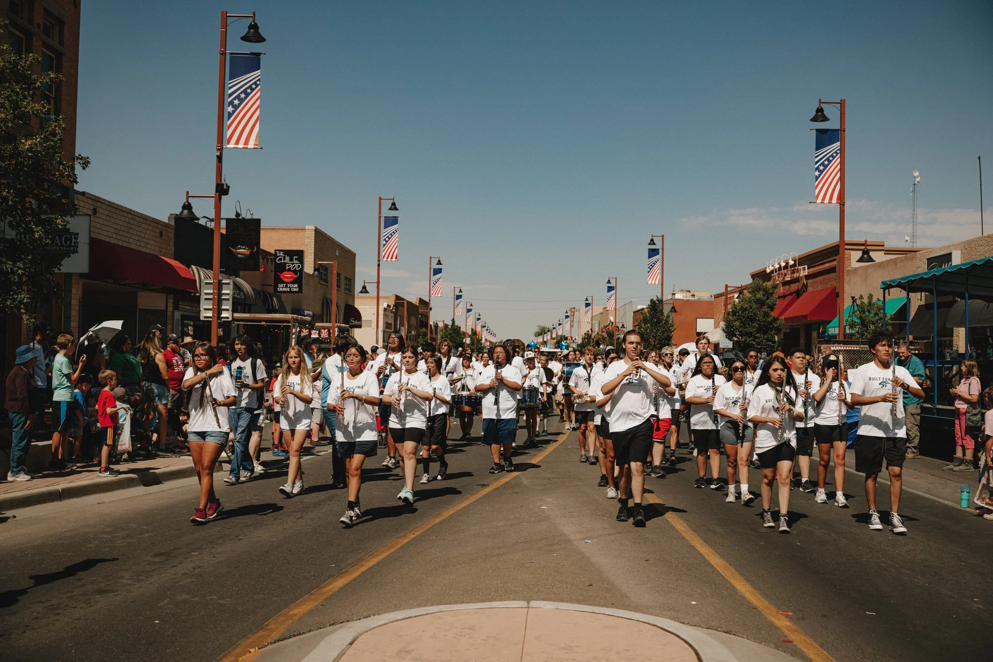 A parade on a city street with a marching band and a large crowd of spectators. The band is wearing white shirts and playing instruments, walking down the street lined with shops and American flags hanging from lampposts.