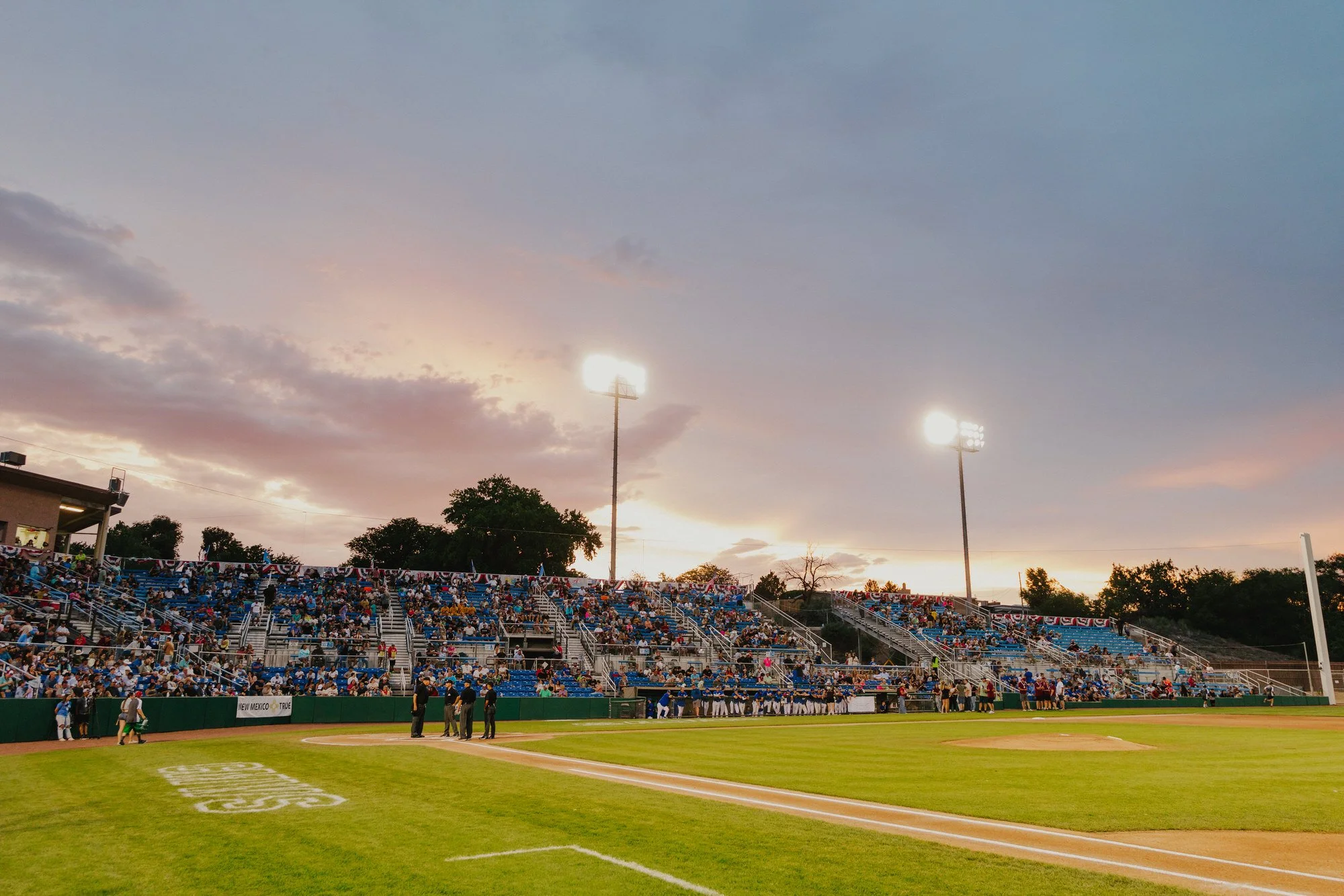 A baseball stadium during evening with spectators in the stands, players on the field, bright stadium lights, and a partly cloudy sky at sunset.