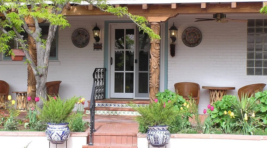 Front porch of a house with a sliding glass door, surrounded by plants and flowers, with patio furniture and decorative elements.
