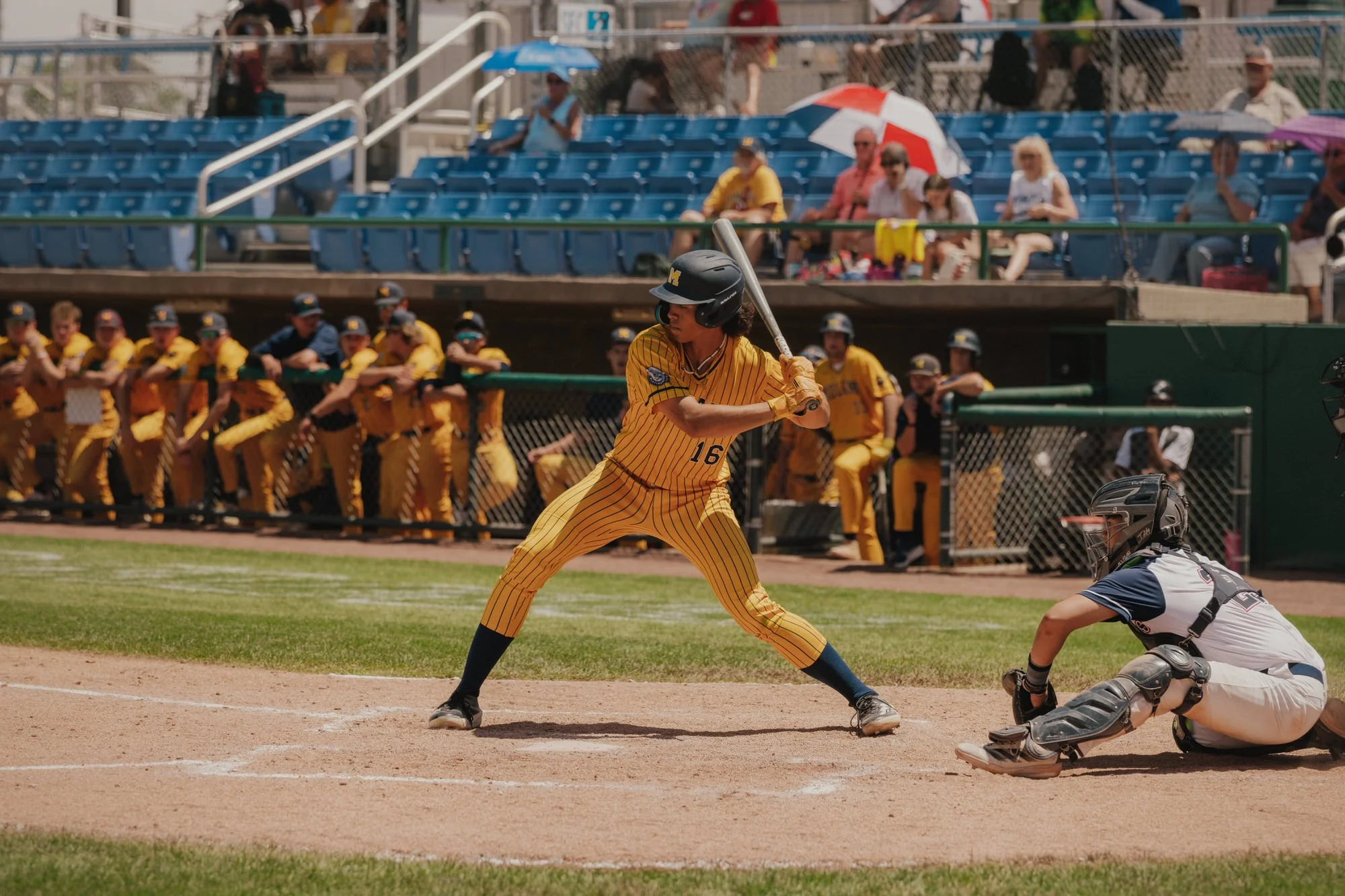 A female baseball player in a yellow striped uniform and black helmet is at bat during a game, with opponents and spectators watching on a sunny day.