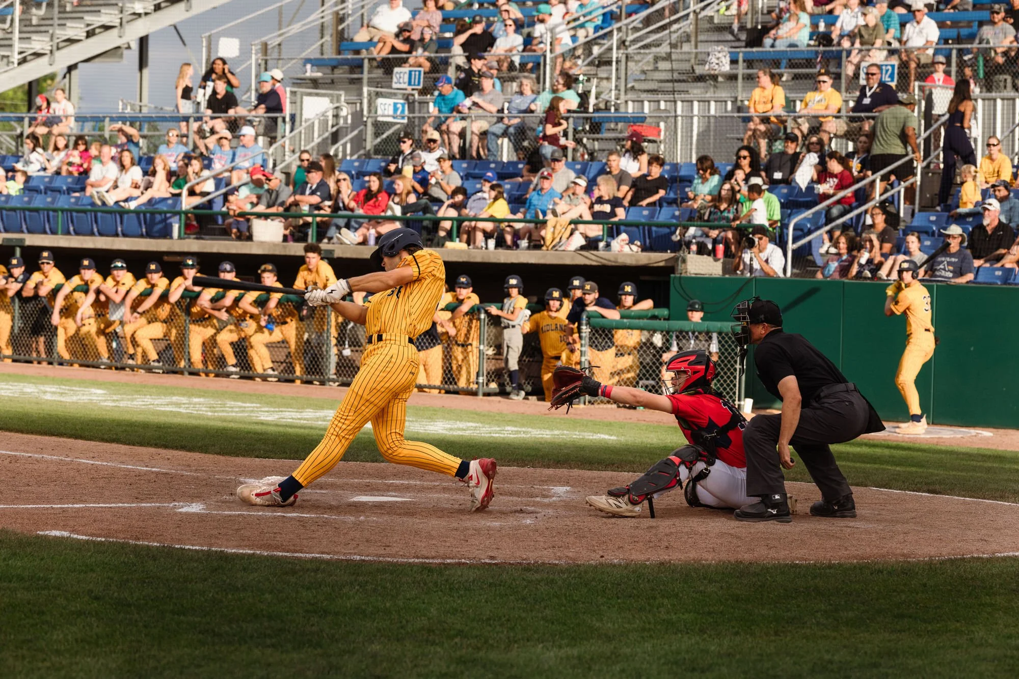 A baseball game in progress showing a batter swinging at a pitch, the catcher and umpire nearby, and spectators watching from the stands.