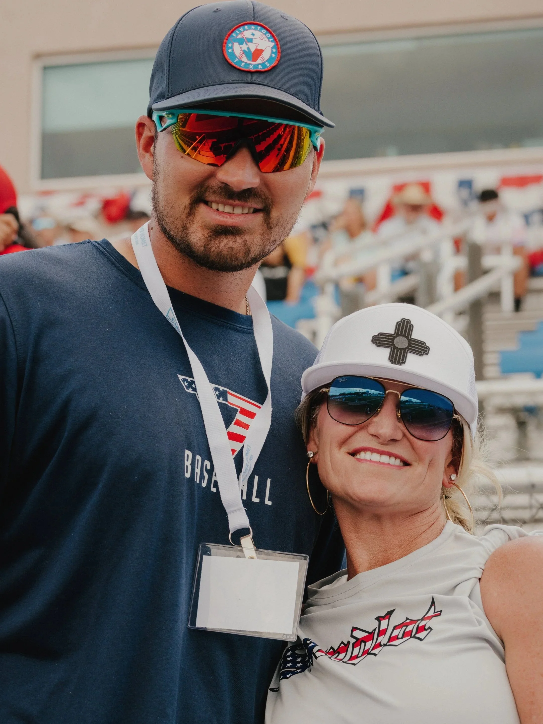 A smiling man and woman at a baseball game. The man is wearing a cap with a logo, reflective sports sunglasses, a blue t-shirt, and a lanyard. The woman is wearing sunglasses, a white cap with a logo, earrings, and a white t-shirt.