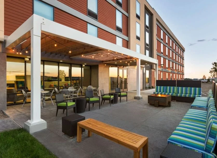 Outdoor terrace of a modern building with seating, tables, string lights, and a fire pit at sunset.