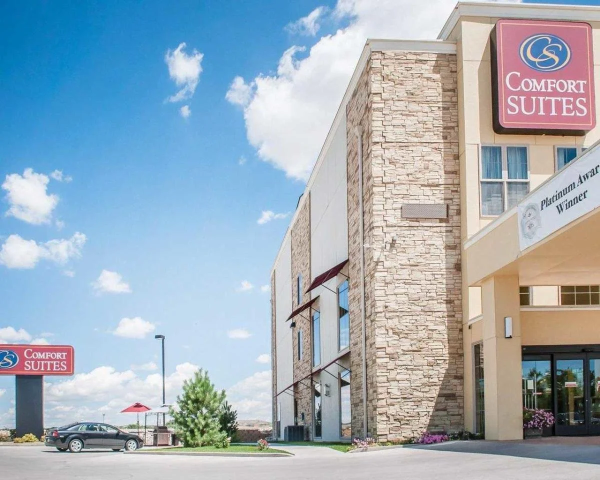 The exterior of a Comfort Suites hotel with a stone wall design, a large pink and blue sign, a parking lot with a car, a small green bush, and a clear blue sky with scattered clouds.
